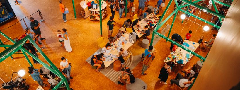 A busy indoor event with people gathered around tables covered in assorted materials. Green metal frames hang above, and various activities are happening throughout the space. Attendees include families and individuals in a creative workshop setting.