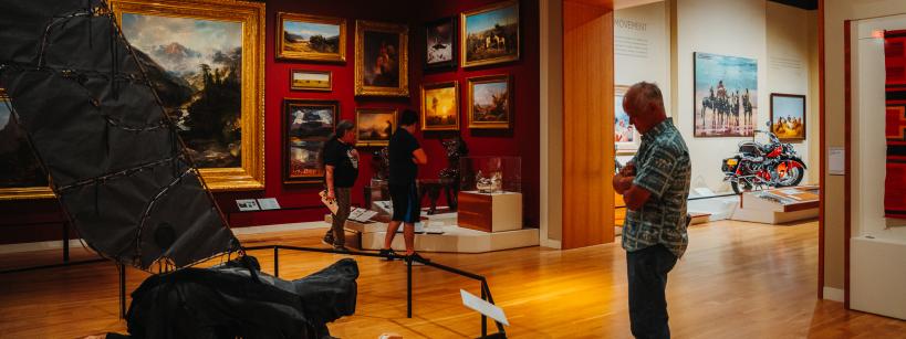 A man stands in an art gallery observing a collection of paintings on red walls. Two other people are viewing exhibits, including a large sculpture and a motorcycle displayed nearby. The room is warmly lit with wooden flooring.