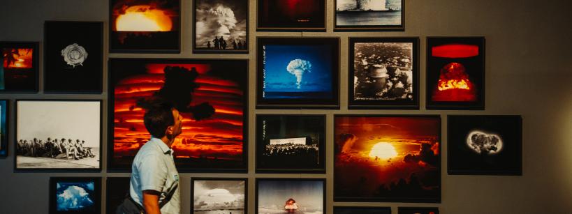 A person observes a gallery wall displaying various framed photographs of nuclear explosions. The images depict dramatic mushroom clouds and fiery skies, capturing the powerful and destructive force of the blasts.