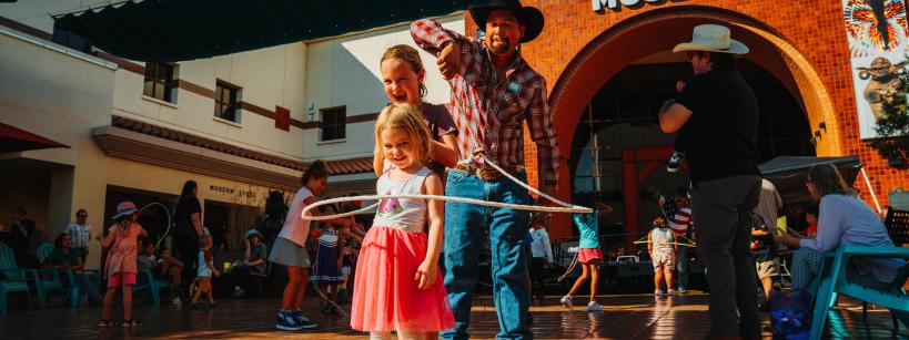 A man helps a young girl hula hoop on a sunlit patio outside a museum. They're surrounded by people, some in cowboy hats. The building in the background has a brick façade with "MUSEUM" written above the entrance.