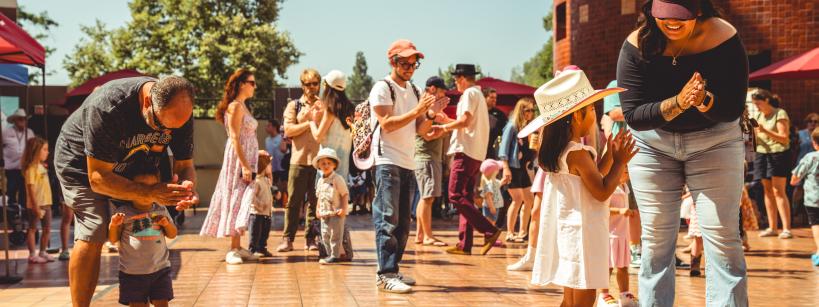 A lively outdoor event with adults and children dancing on a sunlit wooden floor. A woman in a cowboy hat claps along with a young girl, while others enjoy the festive atmosphere. Bright umbrellas and trees frame the background under a clear sky.