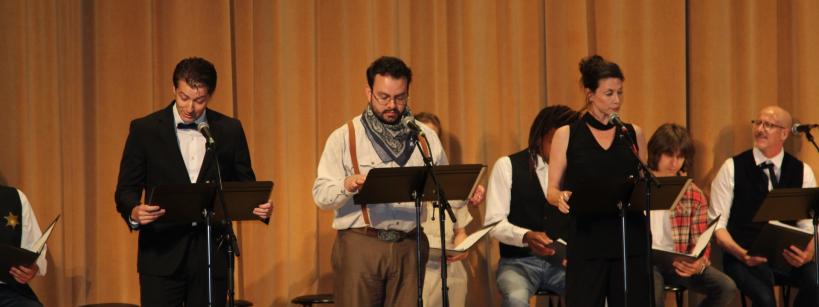 A group of people stand on stage in front of music stands, reading from scripts. They appear to be performing or rehearsing, with some seated in the background. The stage is set against a backdrop of long curtains.