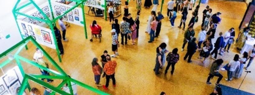 Aerial view of an art gallery with green geometric structures. People browse and converse, examining artworks displayed on the walls. The setting is bright and spacious, creating a lively atmosphere.