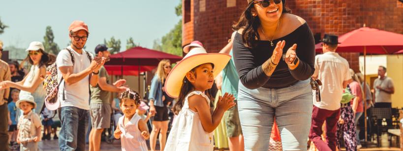 A joyful outdoor scene featuring a group of people dancing and clapping under a sunny sky. A woman wearing sunglasses and a hat claps alongside a young girl in a pink cowboy hat. Other adults and children join the lively gathering.