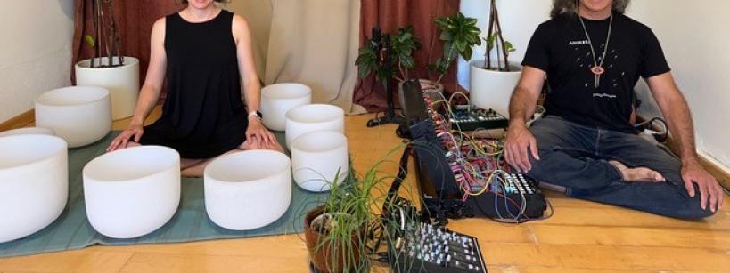 Two people seated on a wooden floor surrounded by sound bowls and electronic equipment. The background features tall green plants and curtains, creating a serene atmosphere. Both individuals are smiling.