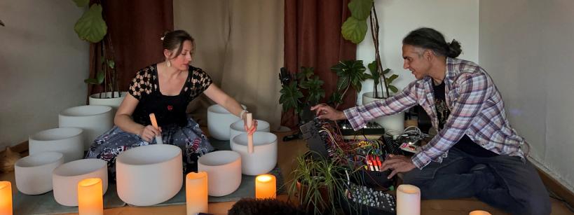 A woman and man seated on the floor playing white crystal singing bowls and electronic synthesizers in a dimly lit room with plants and candles.