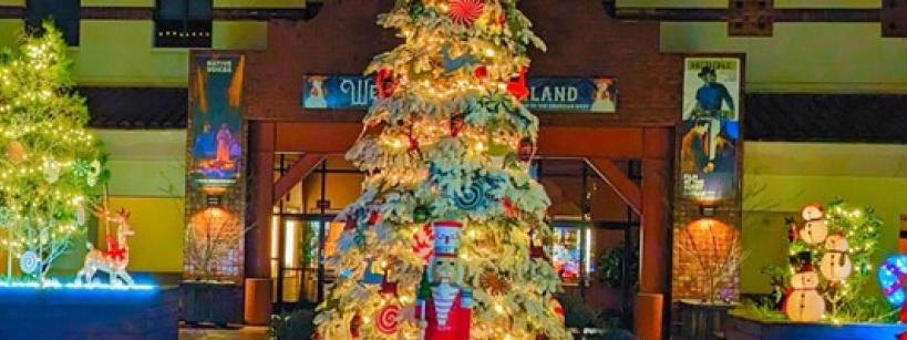 A festive outdoor scene with a large Christmas tree adorned with lights and ornaments in the center. Surrounding decorations include illuminated reindeer, snowmen, and other holiday figures. The setting appears to be a public area at night.