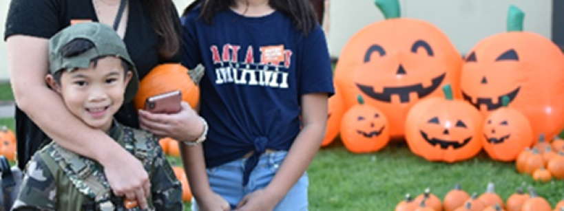 A woman and two children smile in front of Halloween decorations, including large inflatable pumpkins with jack-o'-lantern faces. The boy wears a camouflage outfit, and the girl wears glasses and a casual outfit. A scarecrow stands in the background.