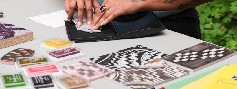 A person with tattoos on their left arm presses a stamp onto an ink pad at a table. Various colorful ink pads and patterned stamps are arranged around them. A lush, green background is visible in the distance.