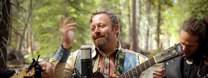 A man with a guitar stands in a forest, smiling as he performs. He is surrounded by two other musicians, one holding a guitar and the other playing a mandolin. A microphone is positioned in front of them. Trees and greenery fill the background.