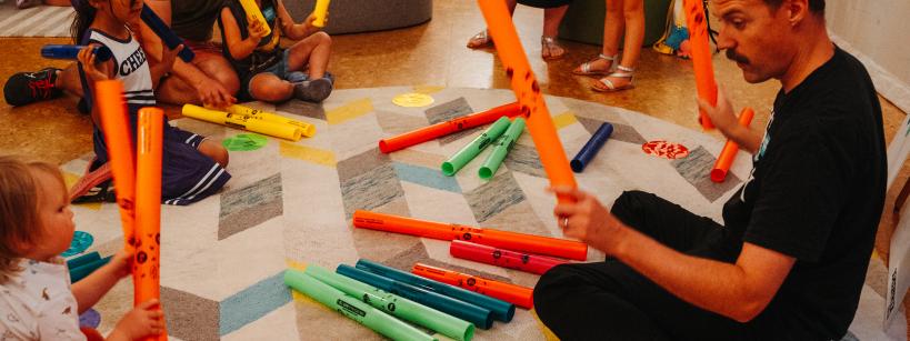 A group of children and adults gather in a colorful play area, engaging with bright music tubes and instruments. They're seated on a patterned rug, actively exploring sounds and rhythms together, creating a lively and interactive atmosphere.