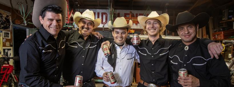 Five men wearing cowboy hats and black shirts with white trim stand in a garage, smiling and holding beer cans. They are surrounded by various tools and equipment, and a motorcycle is partially visible in the background.