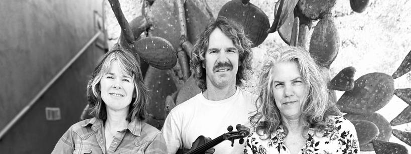 Three people stand in front of a cactus. From left to right, a person holds a guitar, another with a violin, and a third holds a mandolin. All are casually dressed and looking at the camera with neutral expressions. Black and white image.