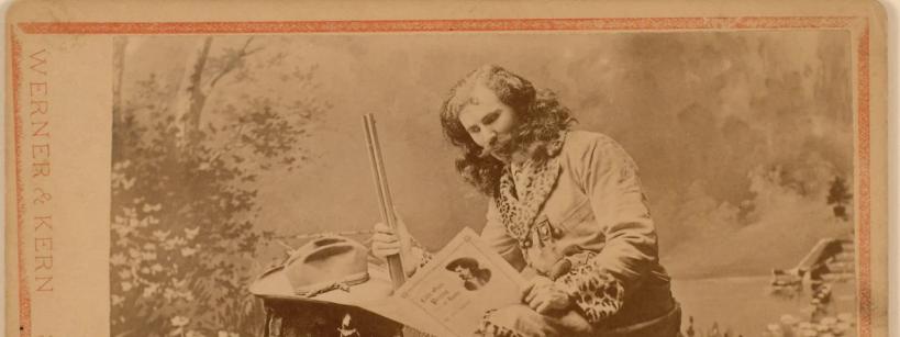A vintage sepia photograph of a person in frontier attire sitting on a tree stump. They hold a rifle and a book while wearing a long coat with fringe details. The background features a painted outdoor scene with trees and foliage.