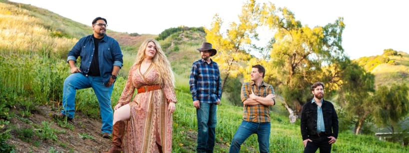 Five people stand on a grassy hillside. The group includes a person in a long dress and boots, and others in casual attire with jeans. One wears a cowboy hat. Trees and rolling hills are in the background under a clear sky.