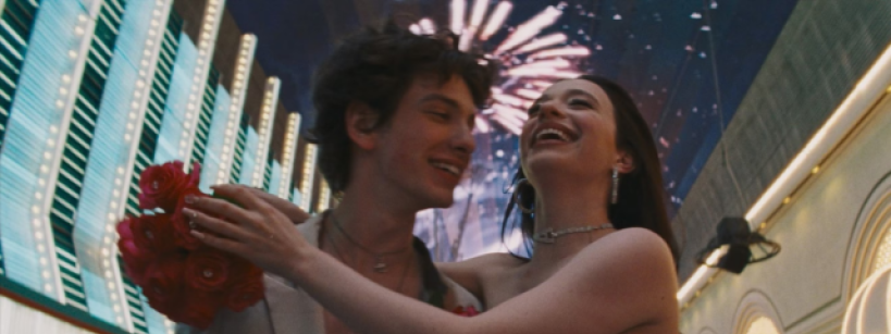 A young couple embracing and smiling joyfully under a lit-up sky filled with fireworks. They are wearing formal attire, with the girl holding a bouquet of red roses. The backdrop includes neon lights and an arcade-like setting, suggesting celebration.