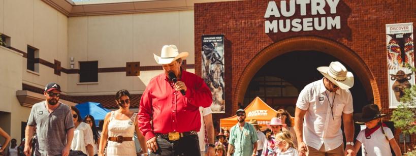 A group of people in cowboy hats and boots are line dancing outside the Autry Museum on a sunny day. The scene is lively, with a mix of adults and children participating. The museum's brick facade is visible in the background.