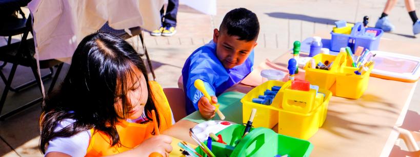 Two children sitting at a table outdoors, wearing aprons and painting. The table is filled with art supplies, including brushes and jars of paint in various colors. The background shows more tables and people.