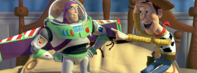 A cowboy toy points excitedly at an astronaut action figure standing on a bed. The bed has a cowboy-themed blanket. The scene takes place in a child's room.