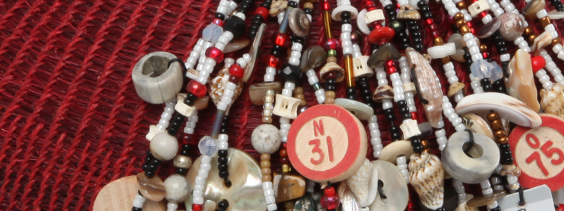 A close-up of numerous colorful beads on a basket with various charms, including circular pieces with numbers "N 31" and "O 75." The necklaces rest on a textured red surface.