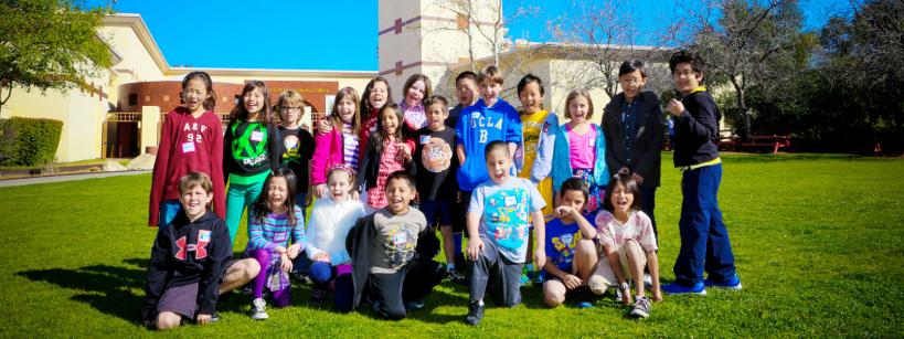 A group of children pose and smile together on a lawn in front of a school building with a tower under a bright blue sky.