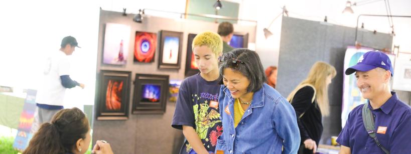 A woman and two teenagers smile and interact with an artist seated at a table at an art fair, while another man stands beside them holding a rolled poster. Colorful artwork is displayed on panels in the background.