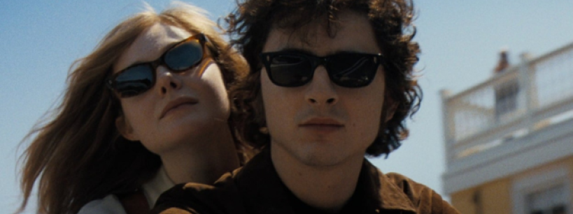 A young woman and man wearing dark sunglasses ride together on a motorcycle under a clear blue sky, with a building in the background. The woman leans on the man's shoulder, both looking calm and composed.