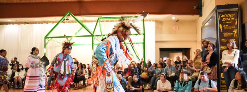 Indigenous dancers in colorful traditional regalia perform in front of a seated and standing audience indoors, with a green structure and exhibits visible in the background.