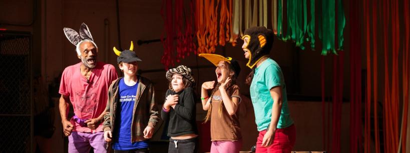 Five people, including children and adults, stand on stage wearing colorful animal hats and costumes. They are smiling and laughing, with colorful streamers hanging in the background.