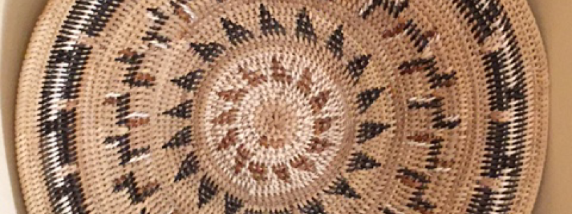 A large, round woven basket with intricate geometric patterns in natural, black, and brown colors is displayed upright against a beige wall on top of a dark wooden surface.