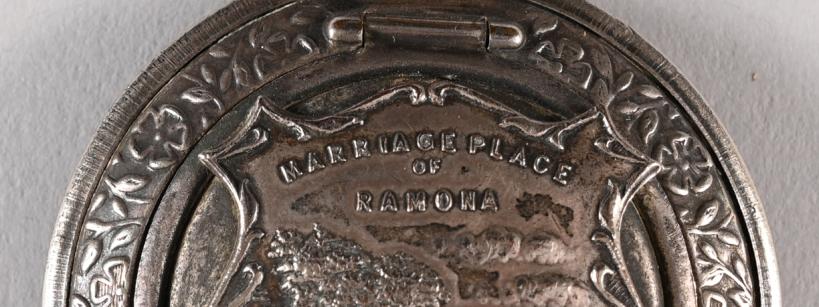 A round silver medallion with an engraved image of a house and trees, inscribed with "Marriage Place of Ramona" and "San Diego, Cal," with decorative patterns around the border and a loop at the top for attachment.