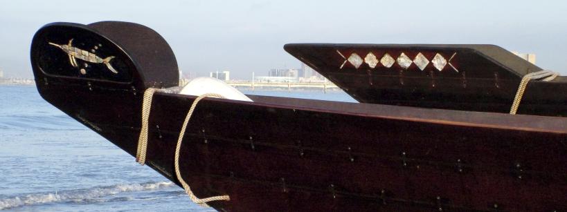 Close-up of the bow of a traditional wooden dhow boat on a beach, with rope detail and geometric decorations; city buildings are visible in the distant background across the water.