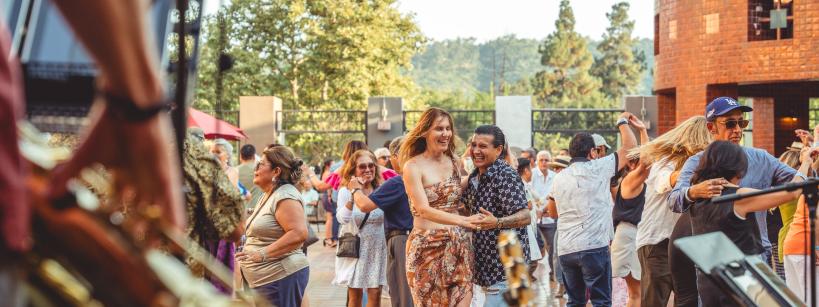 People dance and enjoy live music outdoors on a sunny day. Musicians with instruments are visible in the foreground, and a crowd of smiling dancers fills the background. Trees and brick buildings surround the scene.