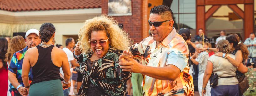 A joyful crowd dances outdoors in front of a building. A smiling woman with curly hair and a man in a colorful shirt are dancing together in the foreground, surrounded by other people enjoying the lively atmosphere.