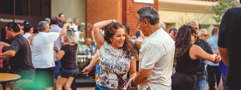 People dancing joyfully in an outdoor plaza, with a band playing in the background. The scene is lively, with men and women smiling and enjoying the music on a sunny day.