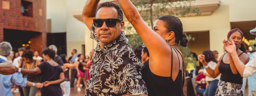 A man and woman dance together in an outdoor courtyard, surrounded by other people who are also dancing. The man wears sunglasses and a floral shirt, while the woman smiles as she spins under his arm.