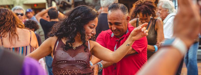 A woman and man dance joyfully outdoors in a crowd, surrounded by people smiling and enjoying the lively atmosphere. The woman raises her arm while the man smiles, closely following her moves.