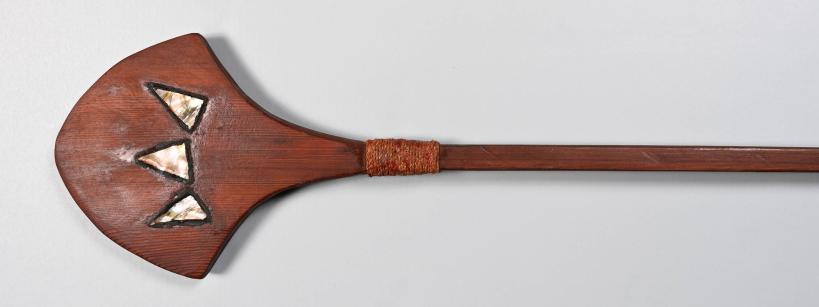 A wooden paddle with a wide, flat blade featuring three triangular mother-of-pearl inlays, and a long, smooth handle wrapped with woven material near the blade.