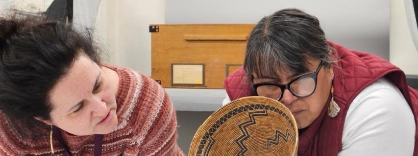 Two women examine a woven basket with intricate patterns; one woman closely inspects the basket while the other leans in with interest. They appear to be in a workspace with papers and objects nearby.
