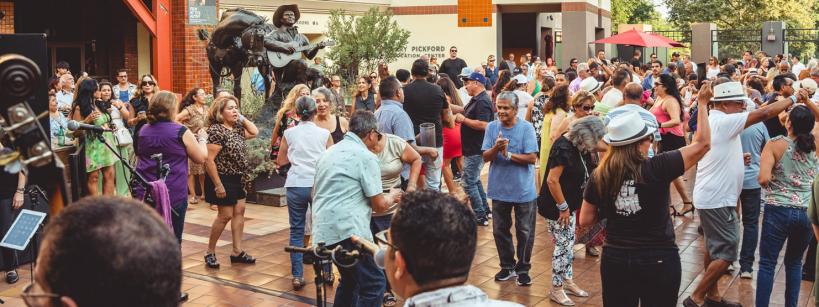 A large group of people dances and socializes outdoors in a plaza, with a musician statue playing guitar in the background and trees and buildings surrounding the lively crowd.
