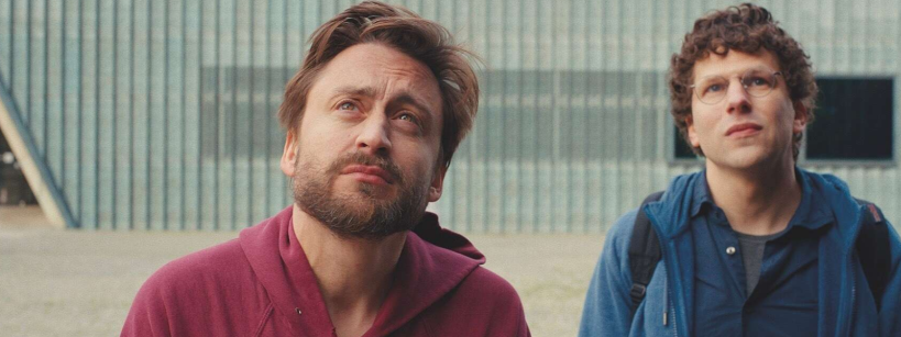 Two men stand outdoors looking upward. One has a beard and wears a red hoodie, while the other has curly hair, glasses, and a blue jacket. The background is a large light blue building with horizontal lines.