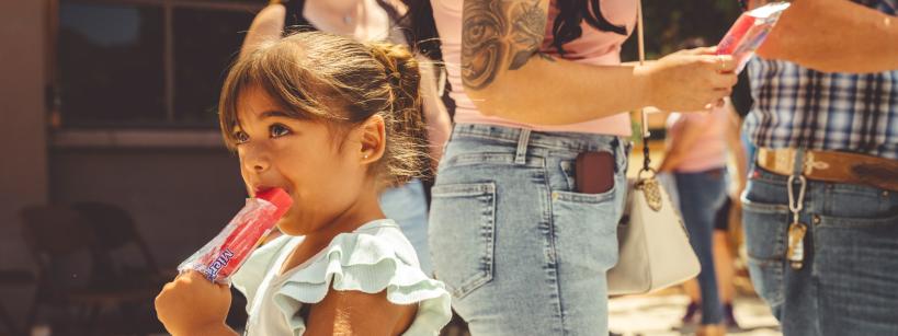 A young girl with brown hair eats a red popsicle while standing outside in a line with adults on a sunny day. She looks off to the side, and people around her are casually dressed in summer clothing.