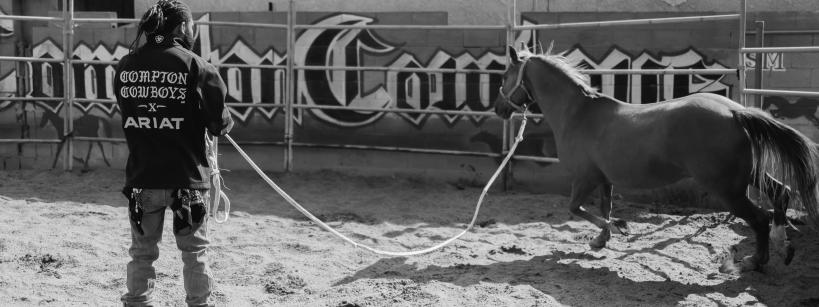 A person wearing chaps and a "Compton Cowboys" shirt holds a lunge line, training a horse in a sandy round pen with a large mural on the wall behind them.