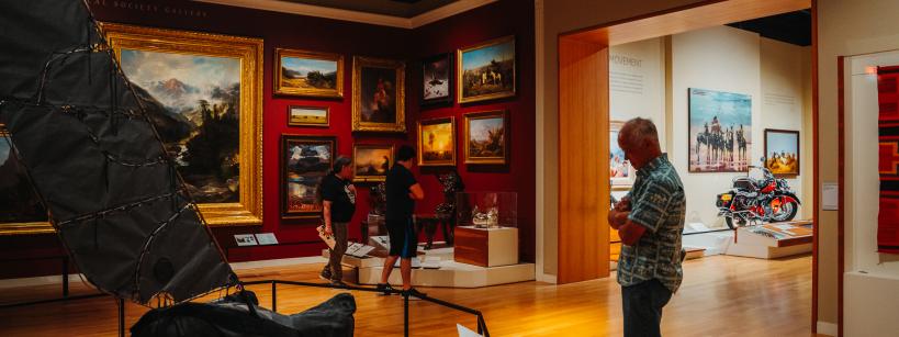 A man observes artworks and exhibits in a museum gallery with red walls, framed paintings, and sculptures. Two other visitors examine displays, and a motorcycle is visible in an adjacent room.