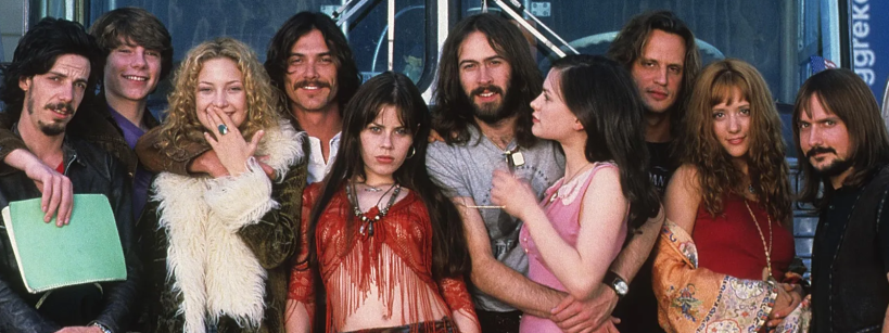 A group of people in 1970s-style clothing pose together, some smiling and some serious, in front of a tour bus. They appear to be members of a rock band and their entourage.