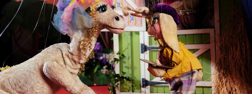 A costumed character resembling a lamb wearing a colorful hat with flowers and veil kneels on a red carpet, facing a smaller puppet in yellow clothes and bunny ears, set in a whimsical indoor scene.