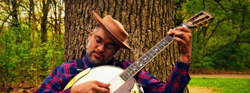 A man wearing a brown hat and glasses sits against a tree in a forest, playing a banjo. He is dressed in a red and blue plaid shirt and appears relaxed and focused on his music.