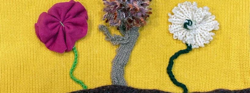 Three textured flowers made from fabric and yarn are attached to a yellow background. The flowers are pink, brown, and white, each with a green stem, standing on a brown horizontal line.