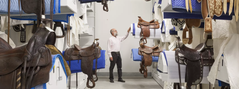 A man stands in a storage room filled with intricately decorated saddles, some hanging and others placed on shelves, surrounded by protective blue and white padding.