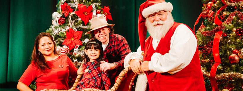 A family and Santa Claus pose together by decorated Christmas trees. The young girl sits in a festive sleigh, smiling, as her parents stand nearby. Santa stands at the front, wearing his classic red suit and hat.
